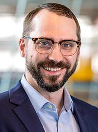 man with beard and glasses smiling at camera wearing a blue button up and navy blazer