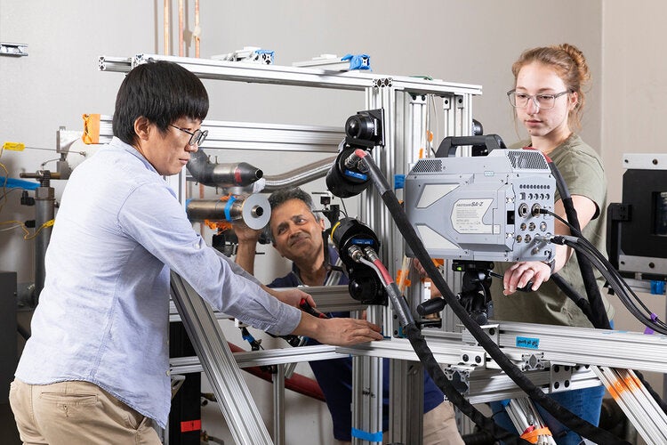 Three researchers examine and adjust components on a metal-framed testing rig, focusing on a mounted sensor and connected cables in a bright lab.