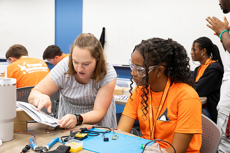 An instructor points to instructions in a workbook while explaining an electronics assembly step to a participant wearing safety glasses.