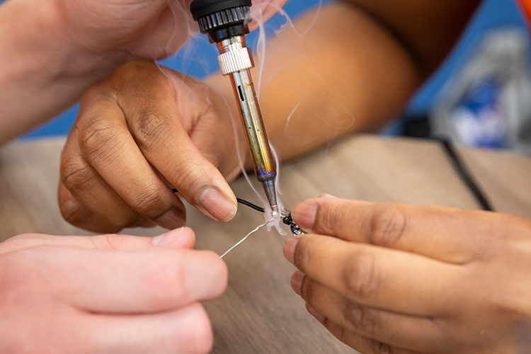 Close-up of several hands using a soldering iron to connect wires to a small electronic circuit board during a classroom electronics project.