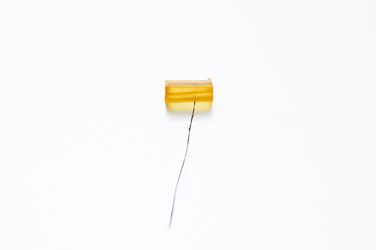 Close-up of a small yellow composite material sample with a thin fiber extending downward, isolated against a plain white background.