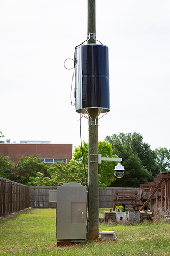 Pole-mounted solar-wrapped device, security-style camera, and open equipment cabinet stand in a fenced outdoor research area.