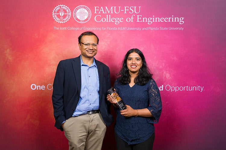 Two individuals stand in front of a FAMU-FSU College of Engineering backdrop, with the woman on the right holding a clear glass award and both smiling at the camera.