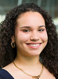 Professional head-and-shoulders portrait of a smiling woman with curly dark hair wearing a navy blue top, gold hoop earrings, and layered necklaces, looking at the camera against a softly blurred indoor background.