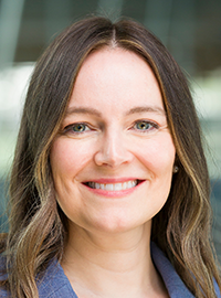 Portrait of a woman wearing a blue blazer, photographed in front of a softly blurred building background.