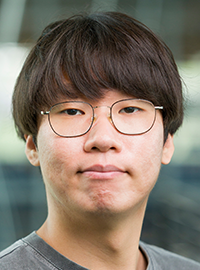 Professional head-and-shoulders portrait of a person wearing round glasses and a gray T-shirt, looking directly at the camera with a neutral expression against a softly blurred indoor background.
