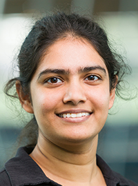 Professional head-and-shoulders portrait of a smiling woman with dark hair pulled back, wearing a black collared knit top, looking directly at the camera against a softly blurred indoor background.