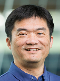 Portrait of a man wearing a blue polo shirt, photographed in front of a softly blurred building background.