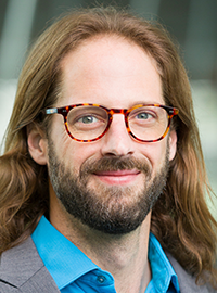 Portrait of a man wearing glasses, a sport coat, and a blue collared shirt, photographed in front of a softly blurred building background.