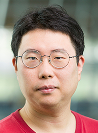 Professional head-and-shoulders portrait of a person wearing round glasses and a red T-shirt, looking directly at the camera with a neutral expression against a softly blurred indoor background.