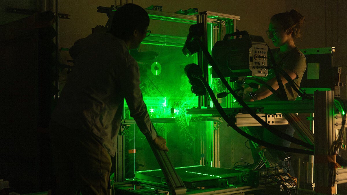 Two researchers adjust a metal-framed experimental rig in a dark lab illuminated by bright green laser light, with visible beams and cables surrounding the apparatus.