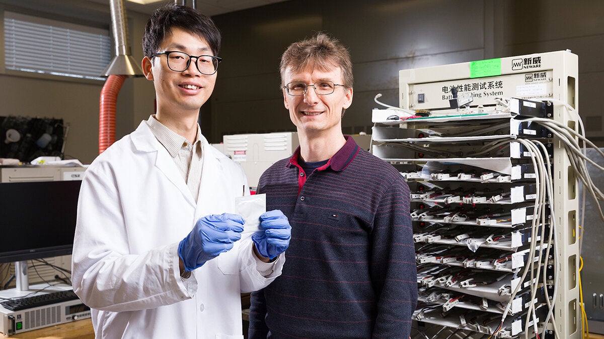 A researcher wearing a lab coat holds a flat, square battery while standing next to a professor in a laboratory.
