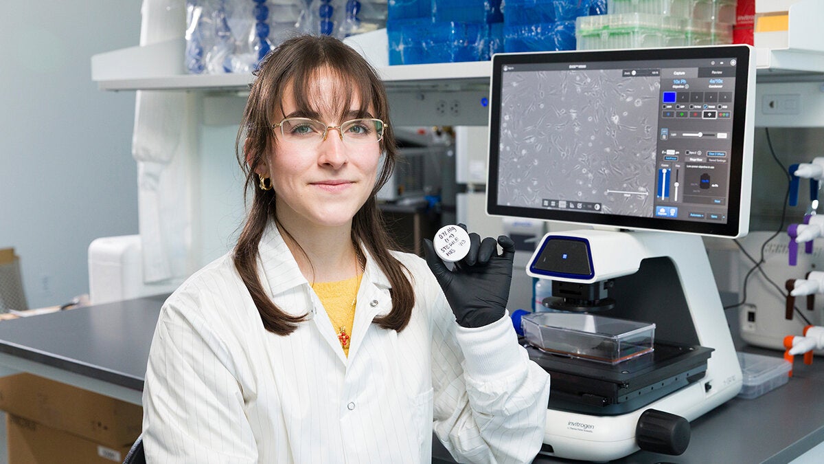 A woman sitting in a lab smiles at the camera in front of a microscope with a screen.