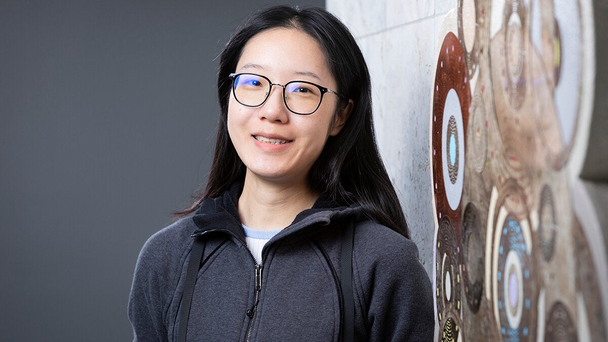Portrait of a woman with long dark hair and glasses leaning beside a large circular patterned wall artwork, arms crossed with a neutral expression.