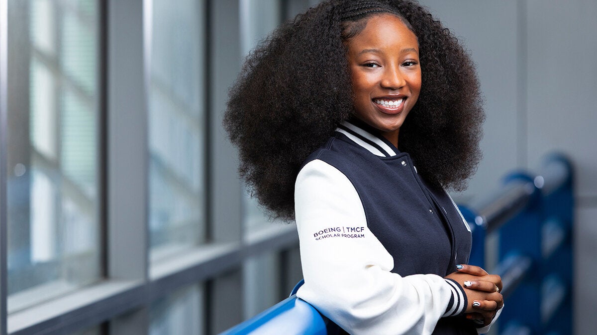 A young woman with curly hair smiles while leaning against a blue railing, wearing a navy and white jacket with Boeing TMFC Scholar Program text on the sleeve.
