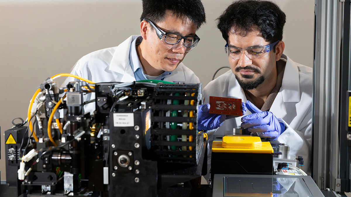 Two researchers wearing lab coats, safety glasses, and gloves examine a laboratory manufacturing machine with electronic equipment and a circuit sample.