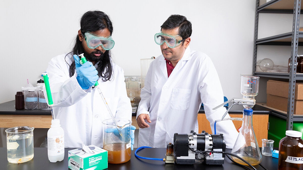 Researcher in safety goggles uses a pipette to transfer liquid into a beaker containing a cloudy brown sample while a colleague observes beside lab equipment.
