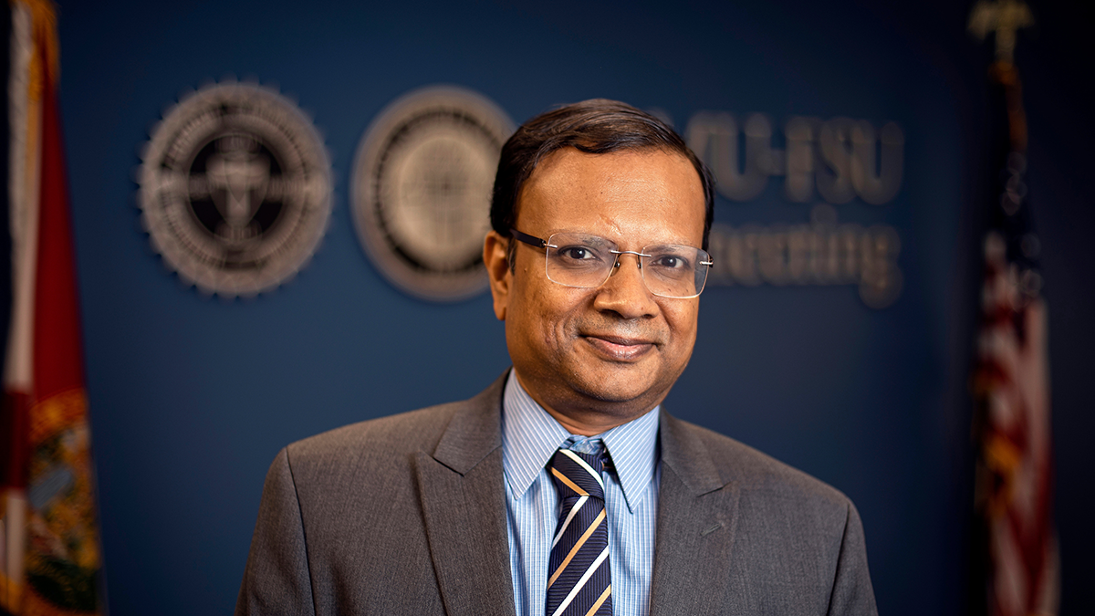 photo of man in glasses blue shirt gray tie smiling against dark background