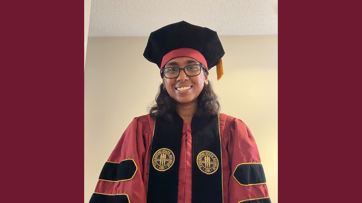 woman in garnet and black phd hood cap gown and black glasses smiling
