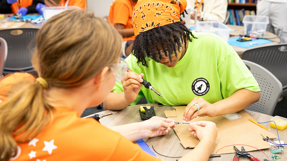 Two students collaborate on an electronics project as one student uses a soldering iron while the other steadies a small circuit board at a classroom worktable.