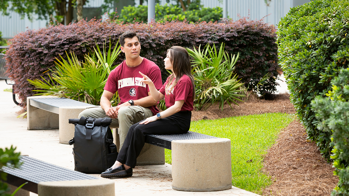 Two students wearing Florida State shirts sit on a bench along a landscaped campus walkway, talking beside a backpack. Outdoor seating, trees, and flowering shrubs surround the area.