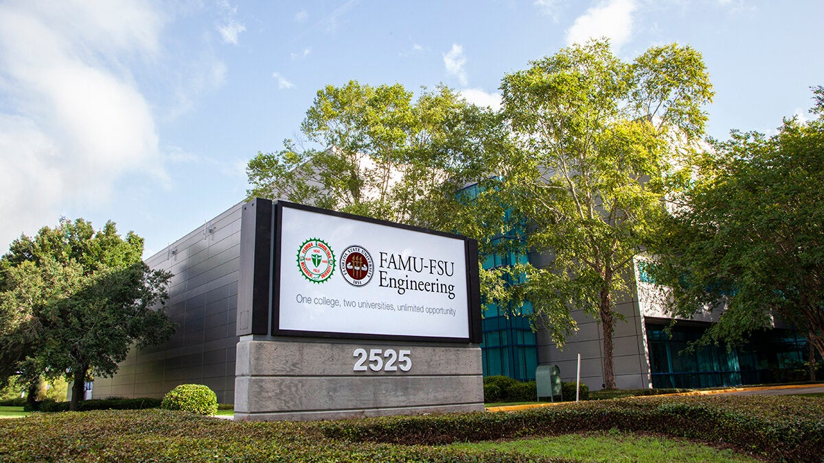 Exterior sign reading "FAMU-FSU Engineering, One college, two universities, unlimited opportunity" outside the college's building at 2525, with trees and a partly cloudy sky in the background.
