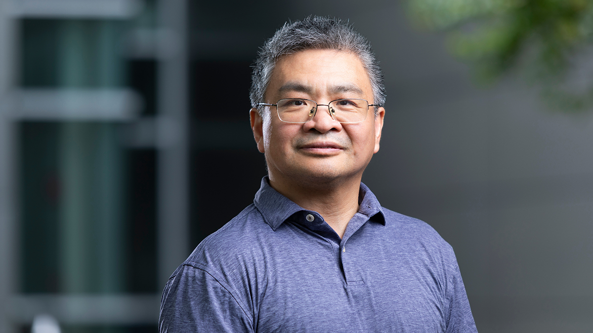 Man wearing glasses and a blue polo shirt stands outdoors against a softly blurred gray building and greenery.