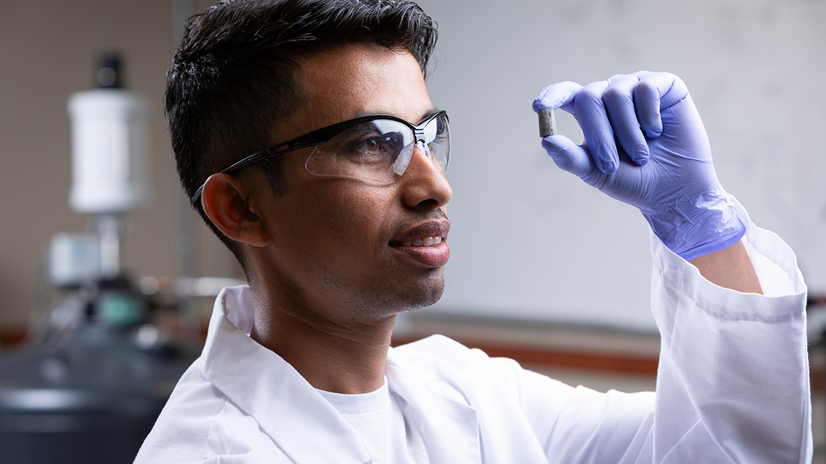 A researcher wearing a lab coat, safety glasses, and a purple nitrile glove examines a small cylindrical sample while working in a laboratory.
