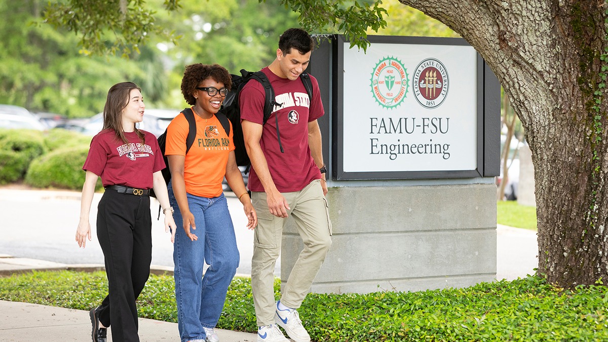 Three students walk together on a campus sidewalk beside a sign reading “FAMU-FSU Engineering.” The students wear university apparel and backpacks while talking beneath a large oak tree.
