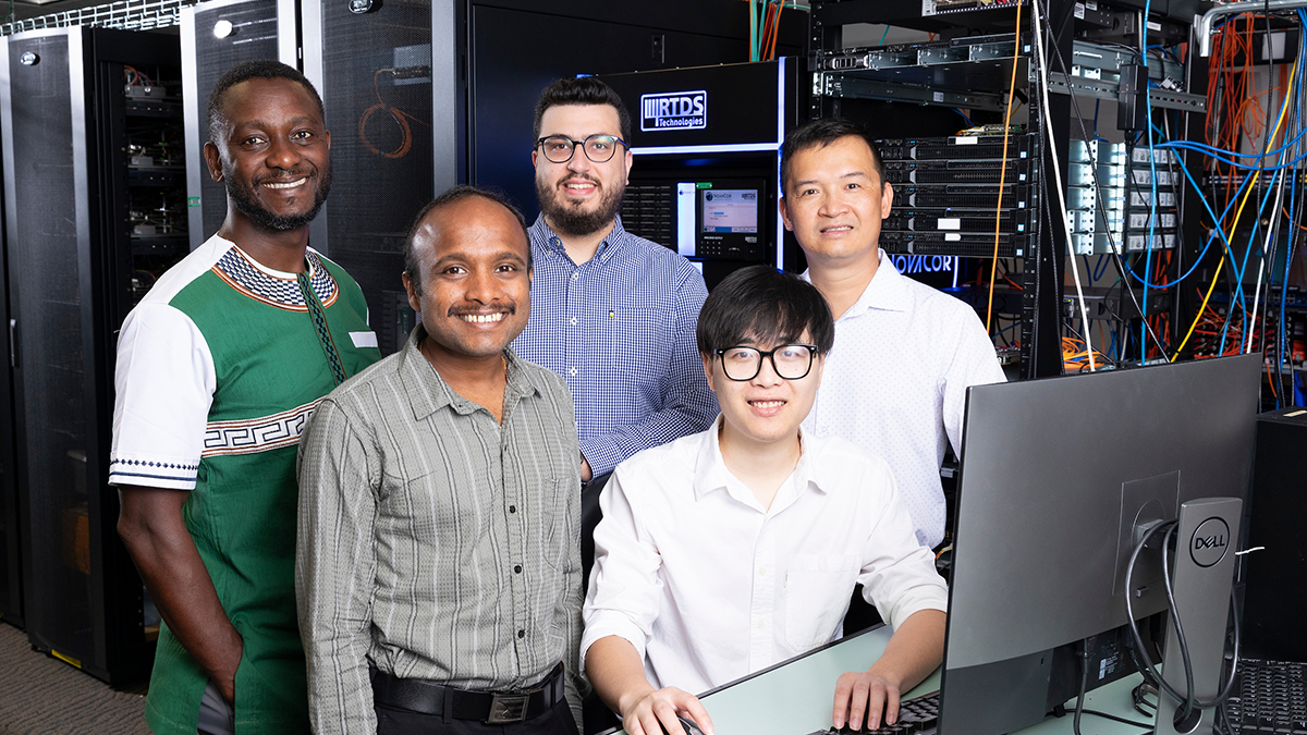 Five researchers stand together in a server room beside a desktop computer, with server racks, networking equipment, and cabling visible in the background.