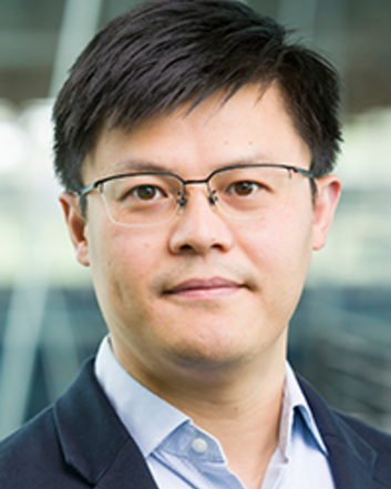 A professional headshot of a man wearing glasses, a navy blazer and a light blue button-down shirt, standing in front of a blurred indoor background.