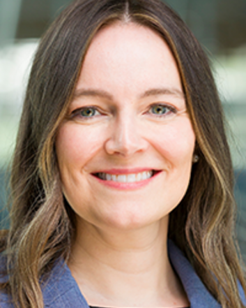 Portrait of a woman wearing a blue blazer, photographed in front of a softly blurred building background.