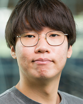 Professional head-and-shoulders portrait of a person wearing round glasses and a gray T-shirt, looking directly at the camera with a neutral expression against a softly blurred indoor background.