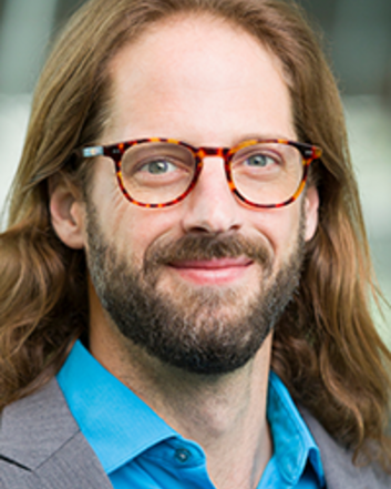 Portrait of a man wearing glasses, a sport coat, and a blue collared shirt, photographed in front of a softly blurred building background.