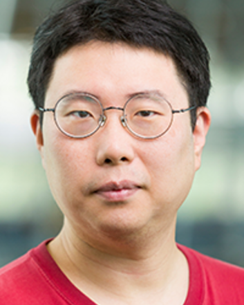Professional head-and-shoulders portrait of a person wearing round glasses and a red T-shirt, looking directly at the camera with a neutral expression against a softly blurred indoor background.