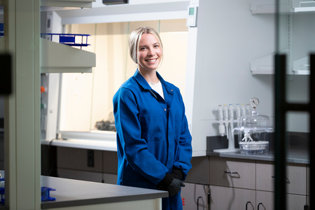 A researcher in a blue lab coat stands in a laboratory near a fume hood and lab equipment.