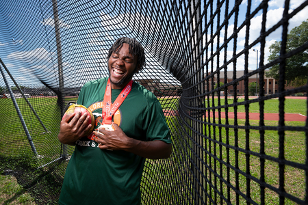 A student athlete laughs while holding a throwing weight and medals inside an outdoor throwing cage