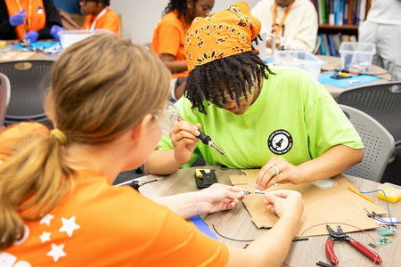 Two students collaborate on an electronics project as one student uses a soldering iron while the other steadies a small circuit board at a classroom worktable.