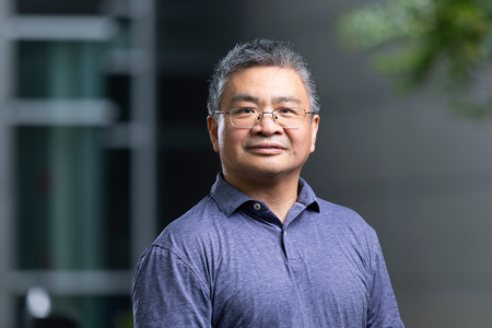 Man wearing glasses and a blue polo shirt stands outdoors against a softly blurred gray building and greenery.