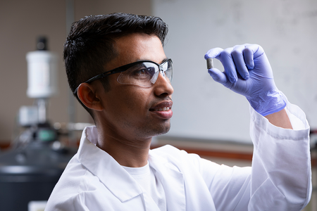 A researcher wearing a lab coat, safety glasses, and a purple nitrile glove examines a small cylindrical sample while working in a laboratory.