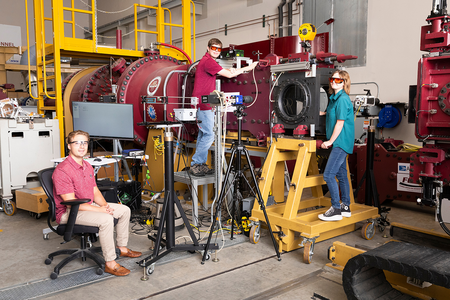 Three students pose around a large red experimental apparatus in an engineering laboratory. One sits at left, one stands on a platform beside the equipment, and one stands on a yellow rolling platform at right.