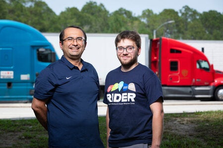 Two men stand outdoors in front of semi-trucks parked side by side, with trees and a clear sky in the background. The man on the right wears a navy RIDER T-shirt.