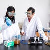 Researcher in safety goggles uses a pipette to transfer liquid into a beaker containing a cloudy brown sample while a colleague observes beside lab equipment.
