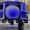 man with braids in black shirt and pants standing next to blue-tinted spacecraft with words rattlers on the rise superimposed on photo