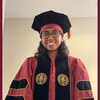 woman in garnet and black phd hood cap gown and black glasses smiling