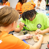 Two students collaborate on an electronics project as one student uses a soldering iron while the other steadies a small circuit board at a classroom worktable.