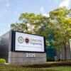 Exterior sign reading "FAMU-FSU Engineering, One college, two universities, unlimited opportunity" outside the college's building at 2525, with trees and a partly cloudy sky in the background.