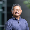 Man wearing glasses and a blue polo shirt stands outdoors against a softly blurred gray building and greenery.