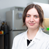 A student wearing a white laboratory coat sits in a research laboratory with scientific equipment visible in the background and smiles at the camera.