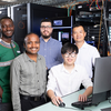 Five researchers stand together in a server room beside a desktop computer, with server racks, networking equipment, and cabling visible in the background.